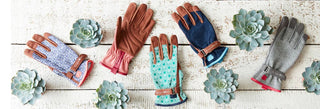Four pairs of winter gloves arranged on a white wooden surface with decorative succulent plants. The gloves include brown leather, navy blue denim, light blue with green floral pattern, and gray with pink lining.