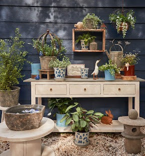A styled outdoor patio scene with a cream console table covered in potted plants, hanging greenery, ceramic pots, a rustic stone bowl, and a dark navy wood plank wall behind it.