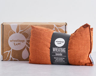 Two orange-brown wheat bags with a black paper label in the center, placed in front of a kraft cardboard box printed with white floral line art and the words 'wheatbags love'.